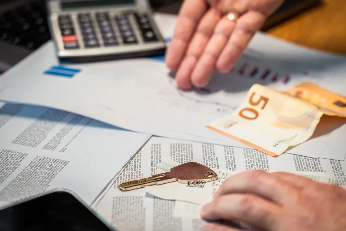 Two hands over printed documents beside a calculator, a 50 euro banknote, and a set of keys.