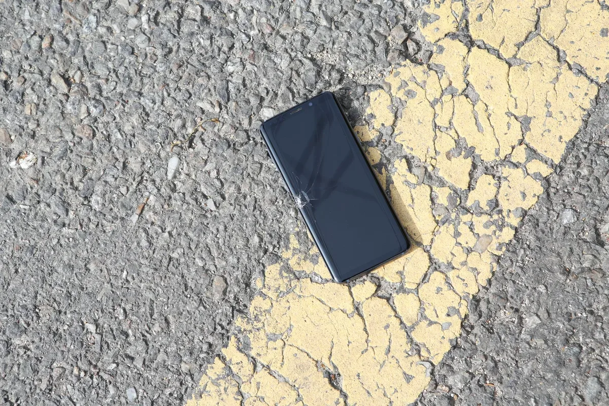 A black smartphone with a cracked screen lies on asphalt beside a worn yellow road line.