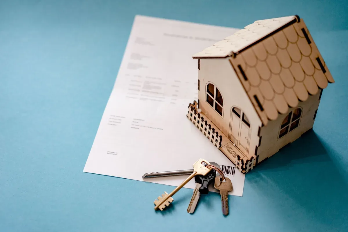 A small wooden model house sits on a printed document beside a set of keys.