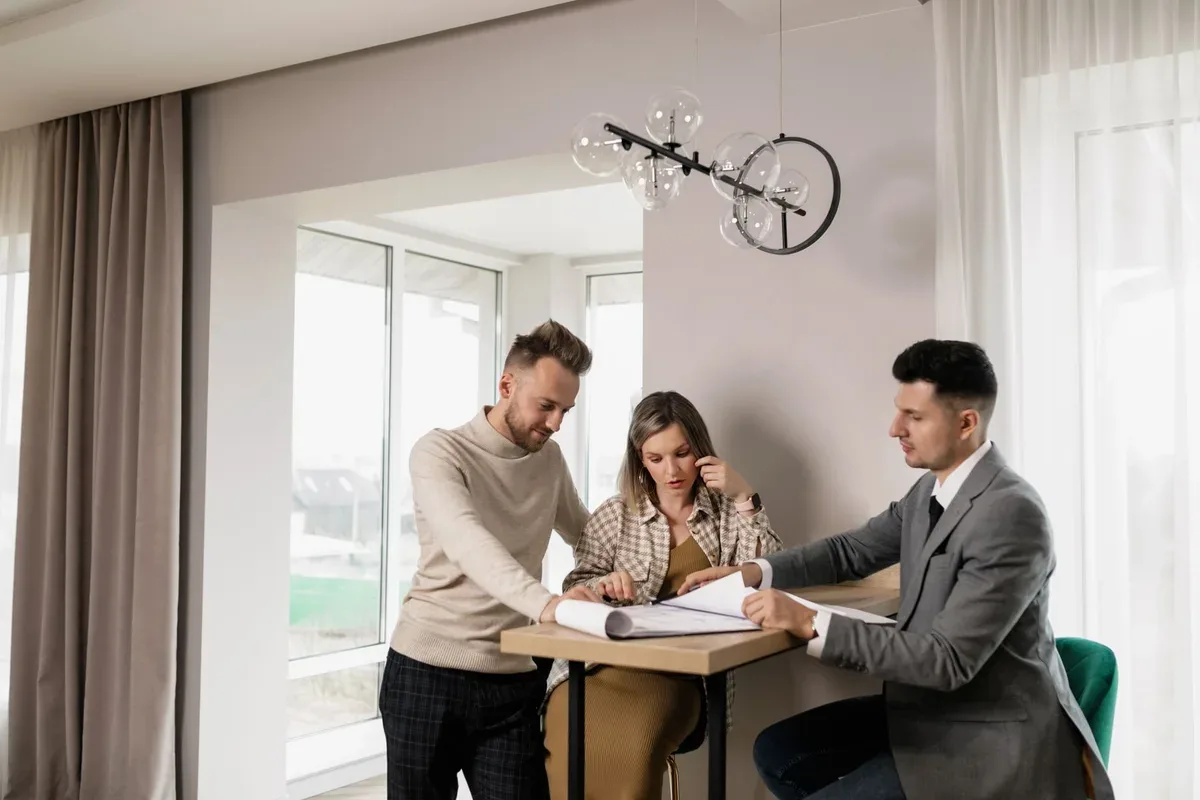 Three people look at an open document on a small table, with one pointing at the pages.